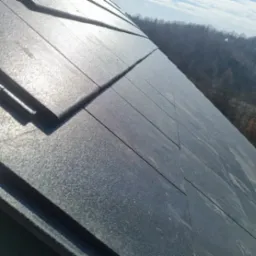 Nowy dach pokryty grafitową dachówką modułową, widok pod kątem w słoneczny dzień, w tle zalesione wzgórza.