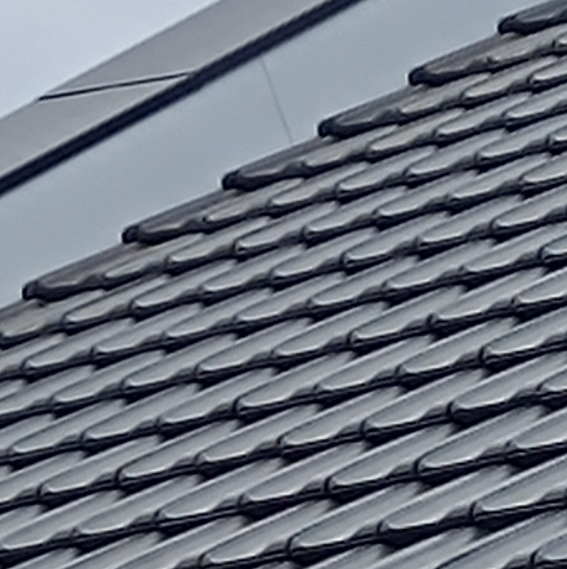 Szare, błyszczące dachówki ceramiczne ułożone ukośnie, widoczne fragmenty obróbek blacharskich w tle.