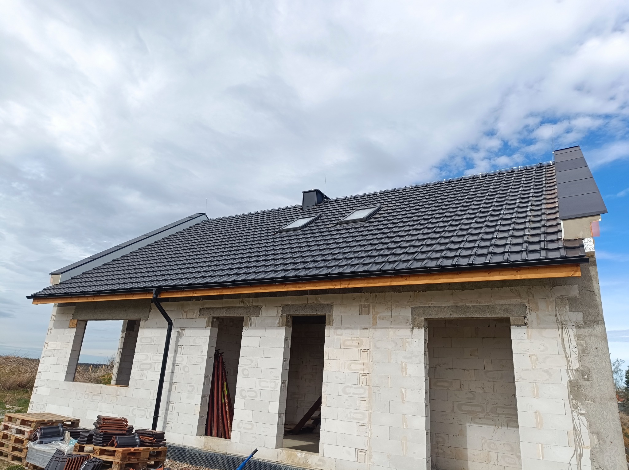 Niedokończony dom z szarej cegły z zamontowanym dachem z ciemnej dachówki, widoczne okna dachowe, rynny i komin, na palecie leżą dachówki.