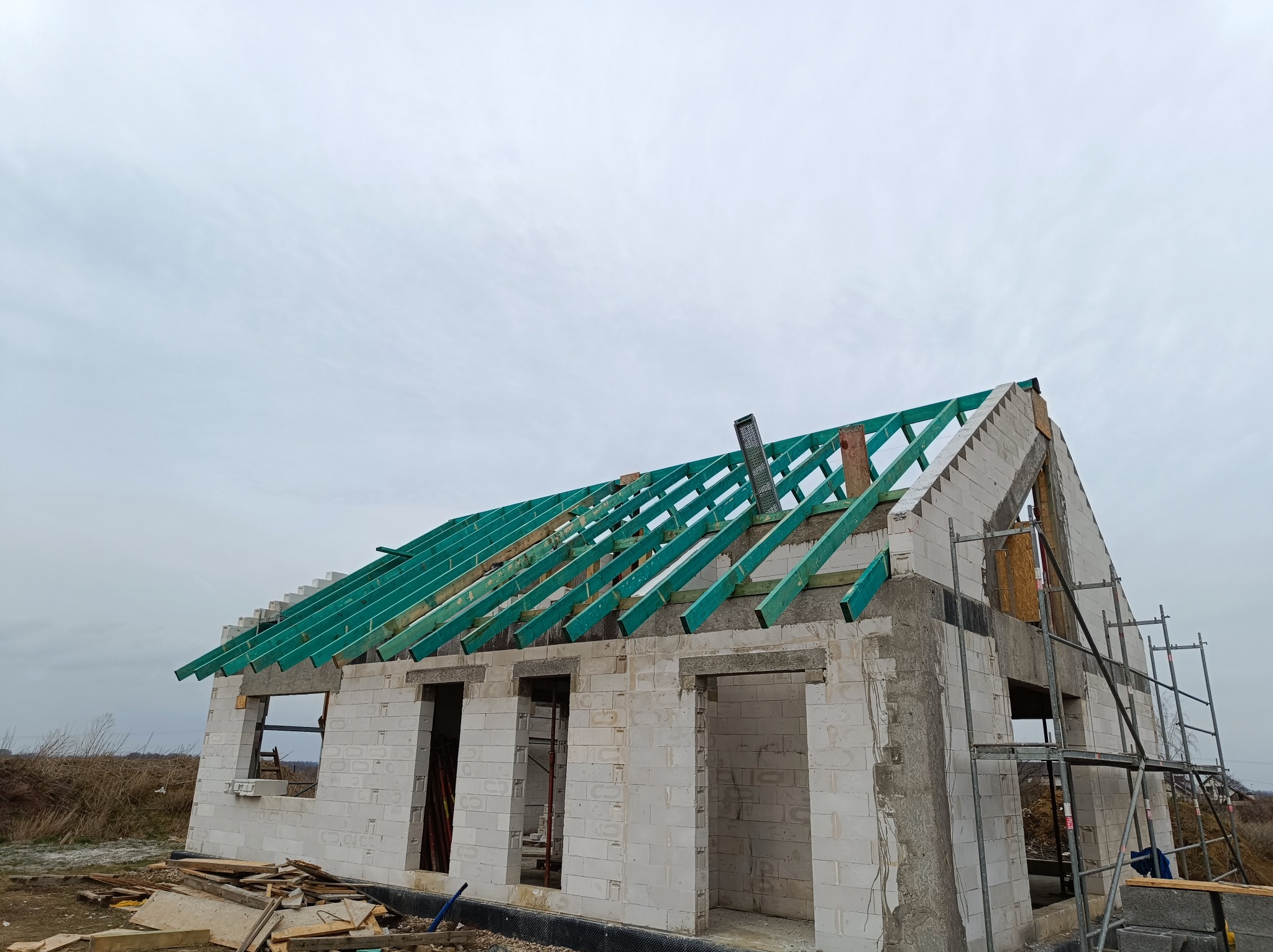 Surowy stan domu jednorodzinnego z bloczków, z widoczną konstrukcją więźby dachowej z impregnowanego drewna w kolorze zielonym, na tle pochmurnego nieba.