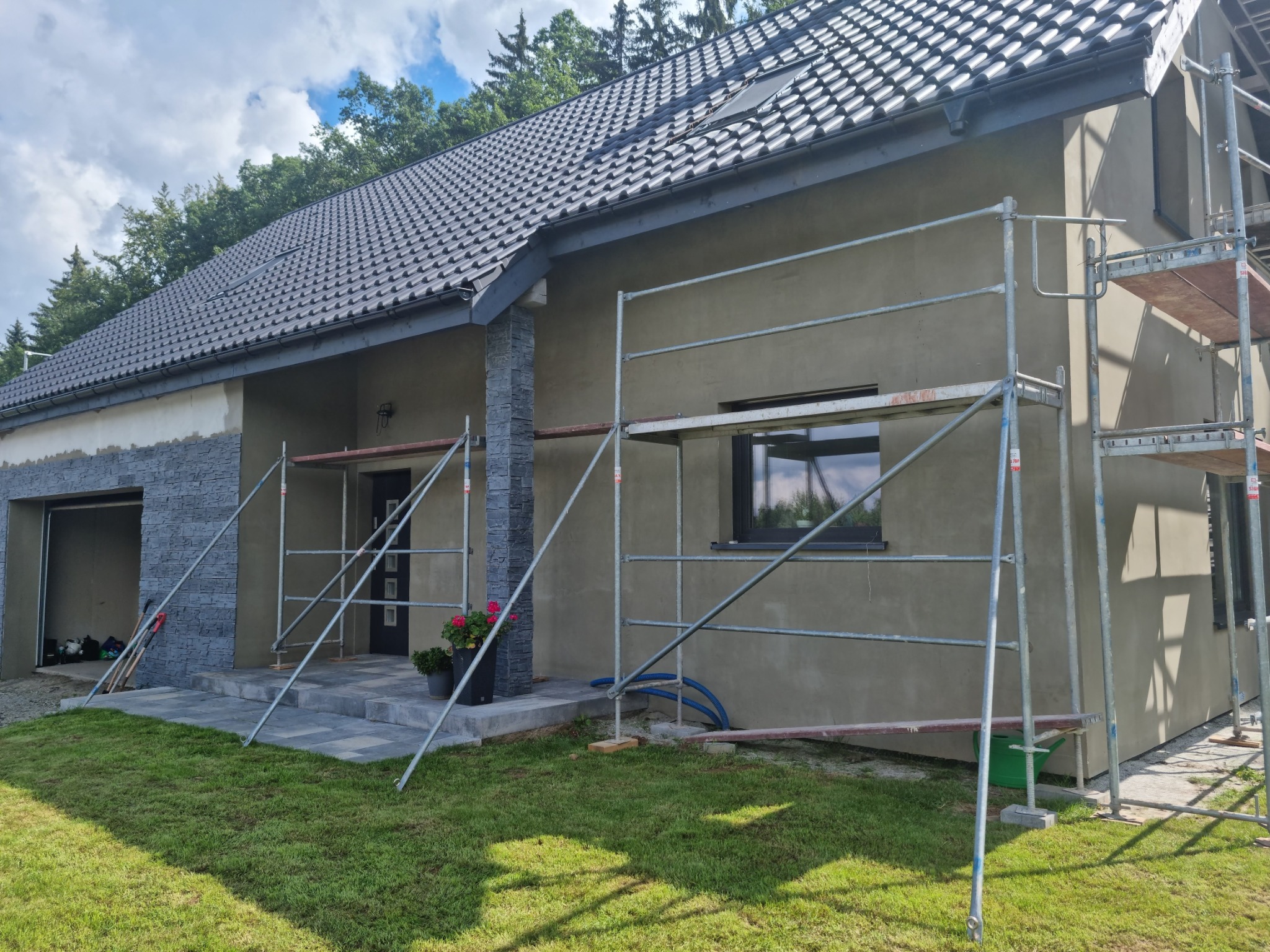 Dom jednorodzinny w trakcie prac wykończeniowych elewacji, widoczne rusztowania, ocieplona ściana w kolorze szarym, fragment wykończony kamieniem elewacyjnym, dachówka ceramiczna w kolorze...