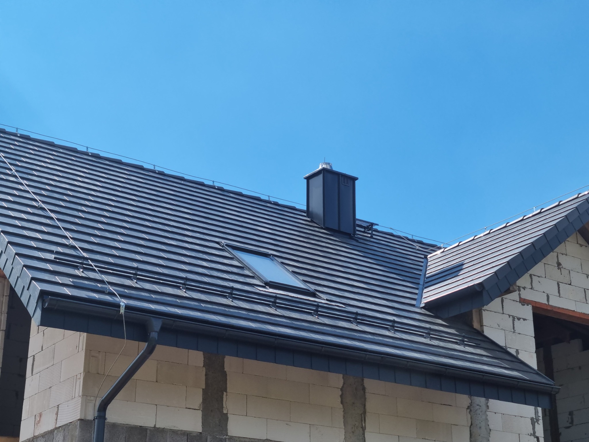 Nowy, grafitowy dach z dachówki ceramicznej z oknem połaciowym i kominem, na budynku w trakcie budowy z białego bloczka, na tle błękitnego nieba.