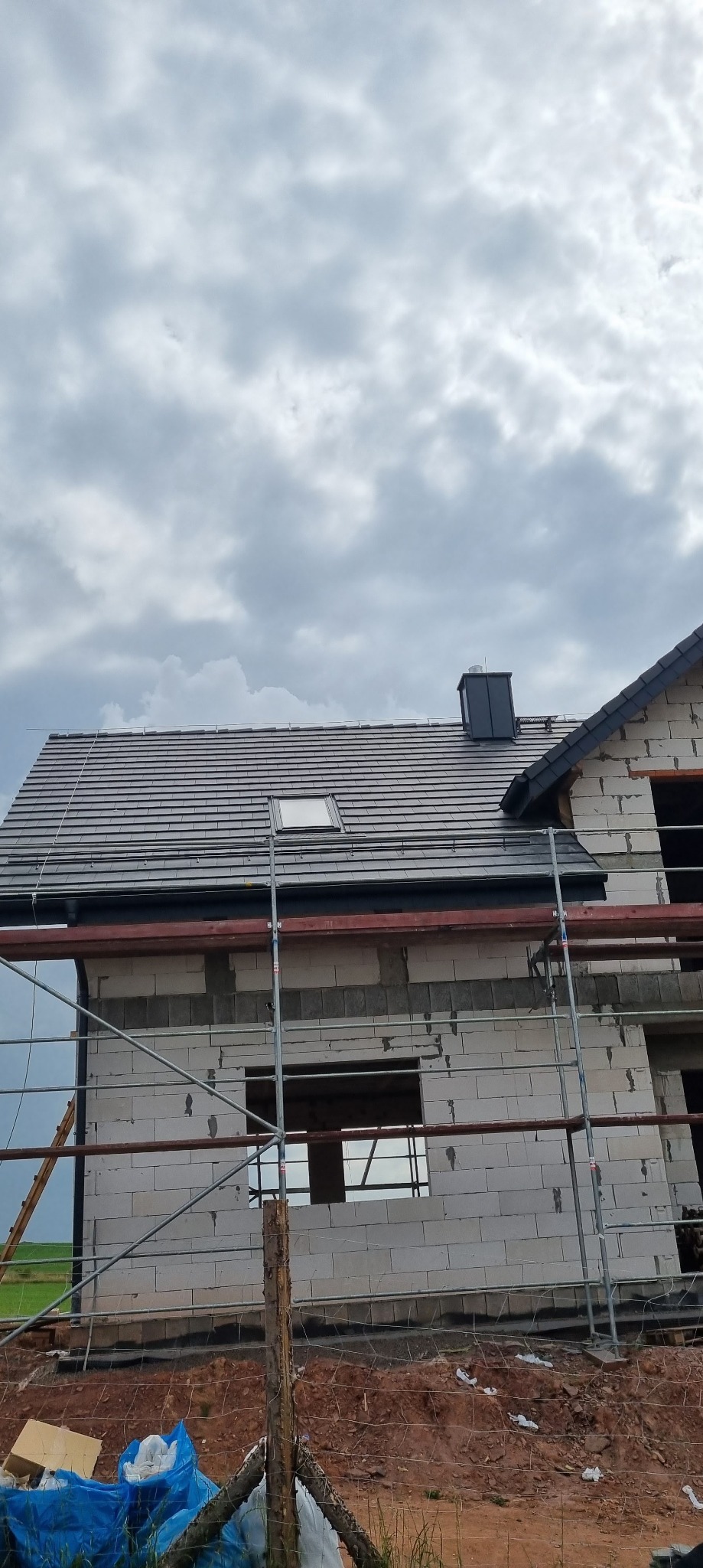 Dom w trakcie budowy z szarą dachówką, widoczne rusztowanie i fragment elewacji z pustaków.