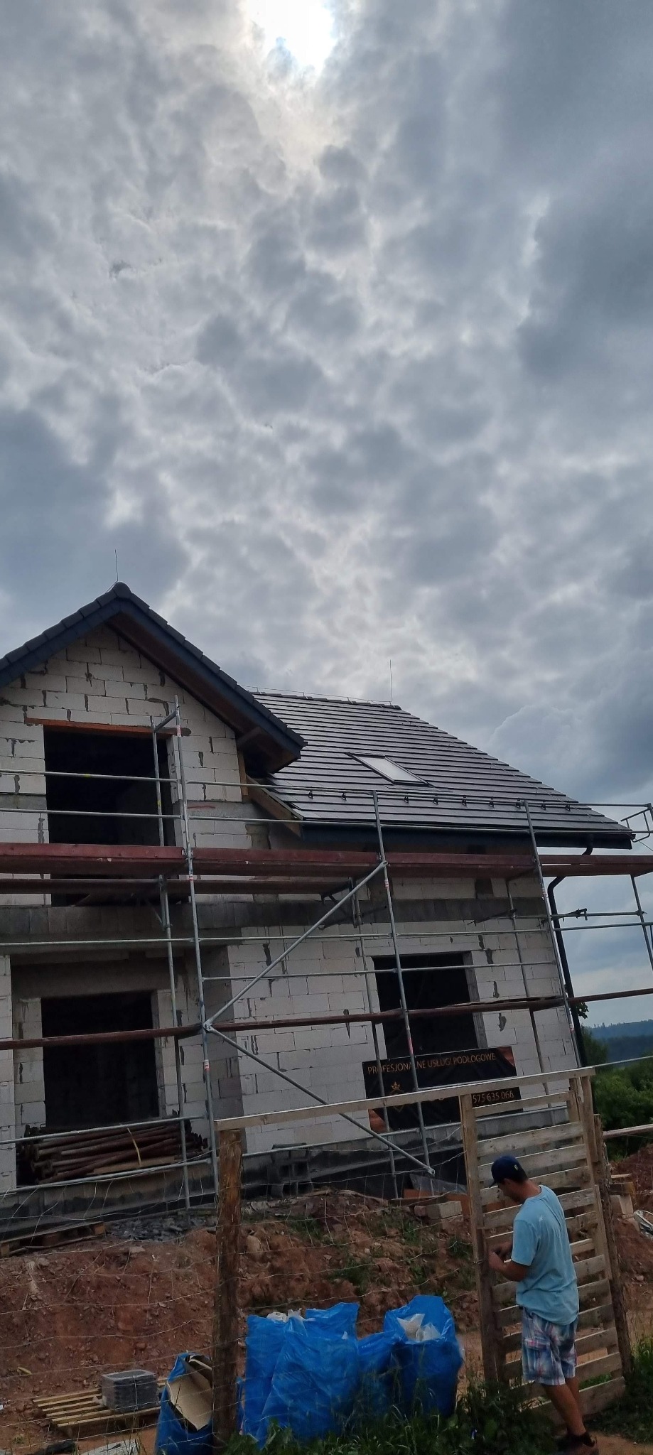Dom w trakcie budowy z częściowo ukończonym dachem z dachówką, widoczne rusztowania i pracownik przy ogrodzeniu, pochmurne niebo w tle.