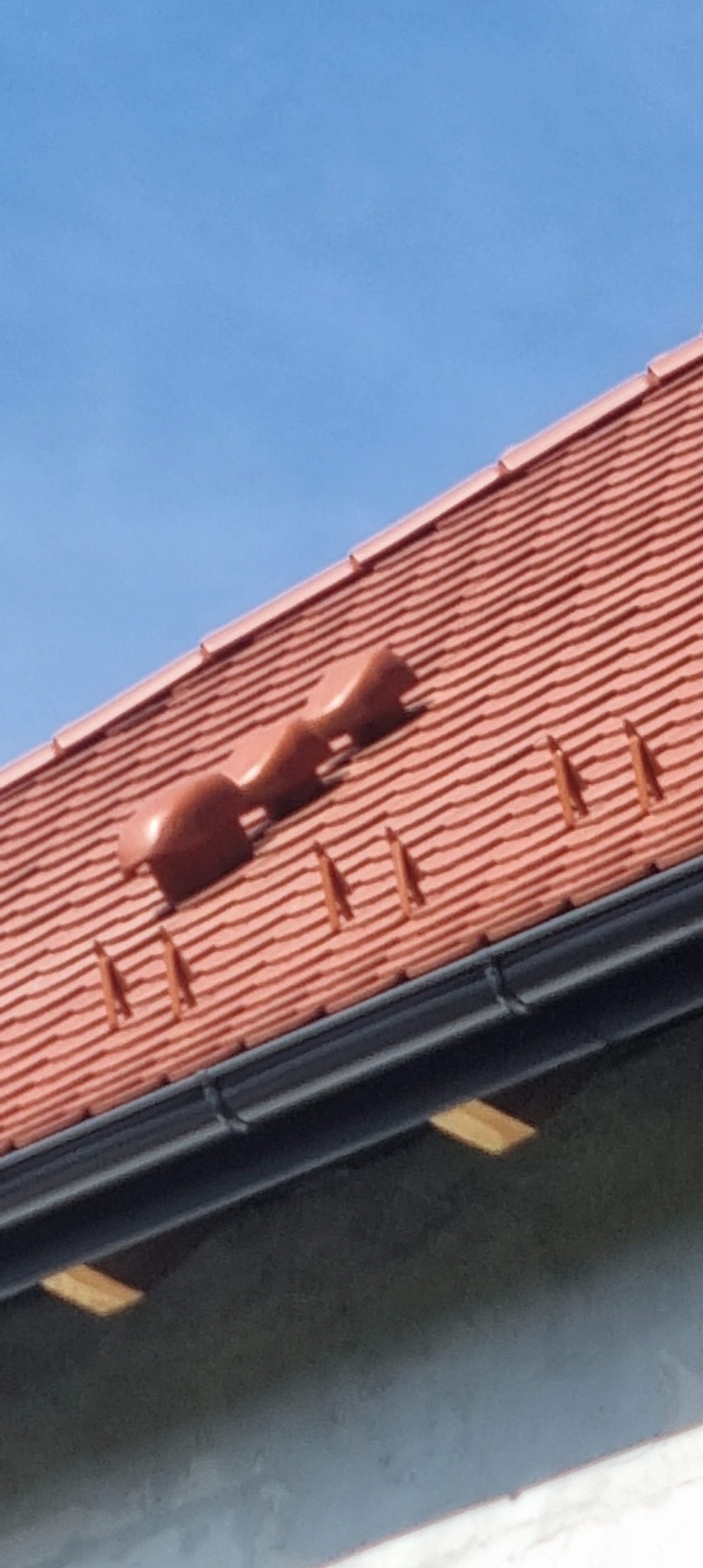 Czerwona dachówka ceramiczna z odpowietrznikami i rynną w kolorze czarnym, widok z dołu na tle błękitnego nieba.