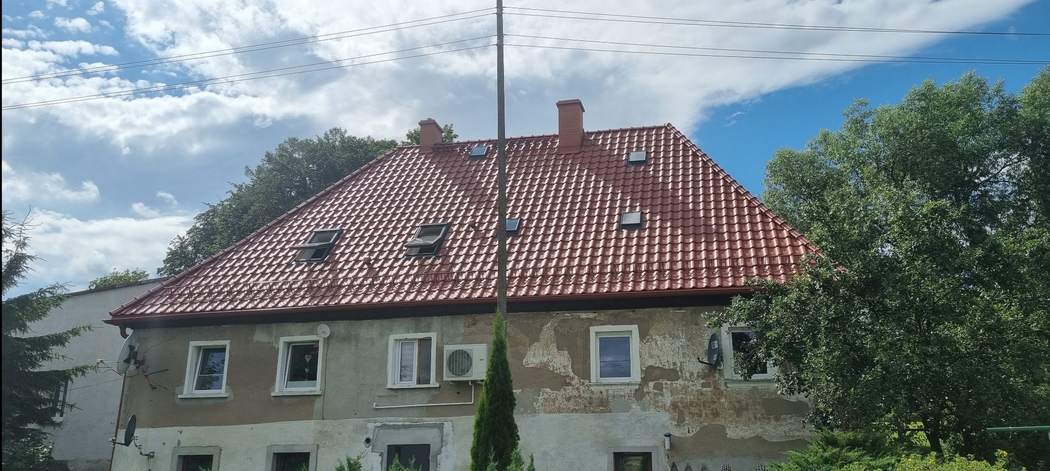 Czerwony dach z dachówkami na dwupiętrowym domu z otwartymi oknami dachowymi i kominami, widok z dołu na tle nieba z chmurami.