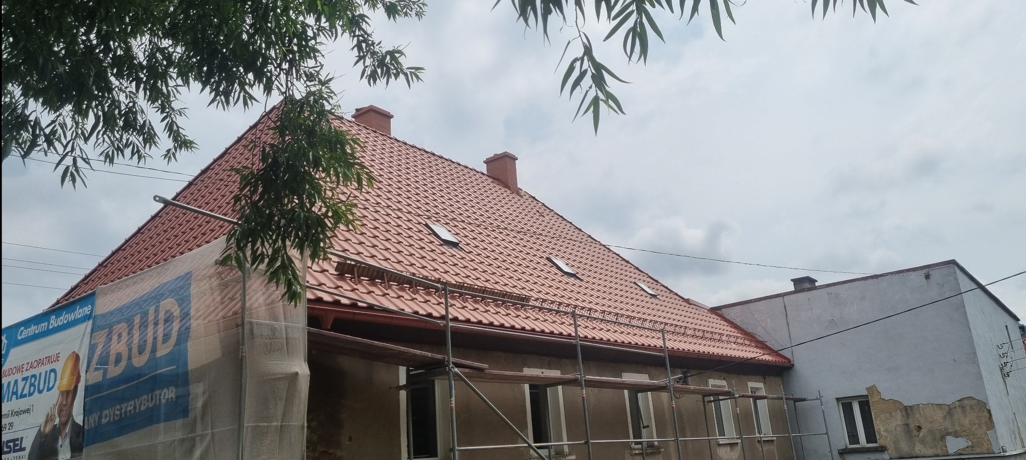 Dom z nowym, czerwonym dachem z dachówki, widoczne okna dachowe i kominy, rusztowanie częściowo zasłonięte banerem reklamowym, ujęcie z dołu.