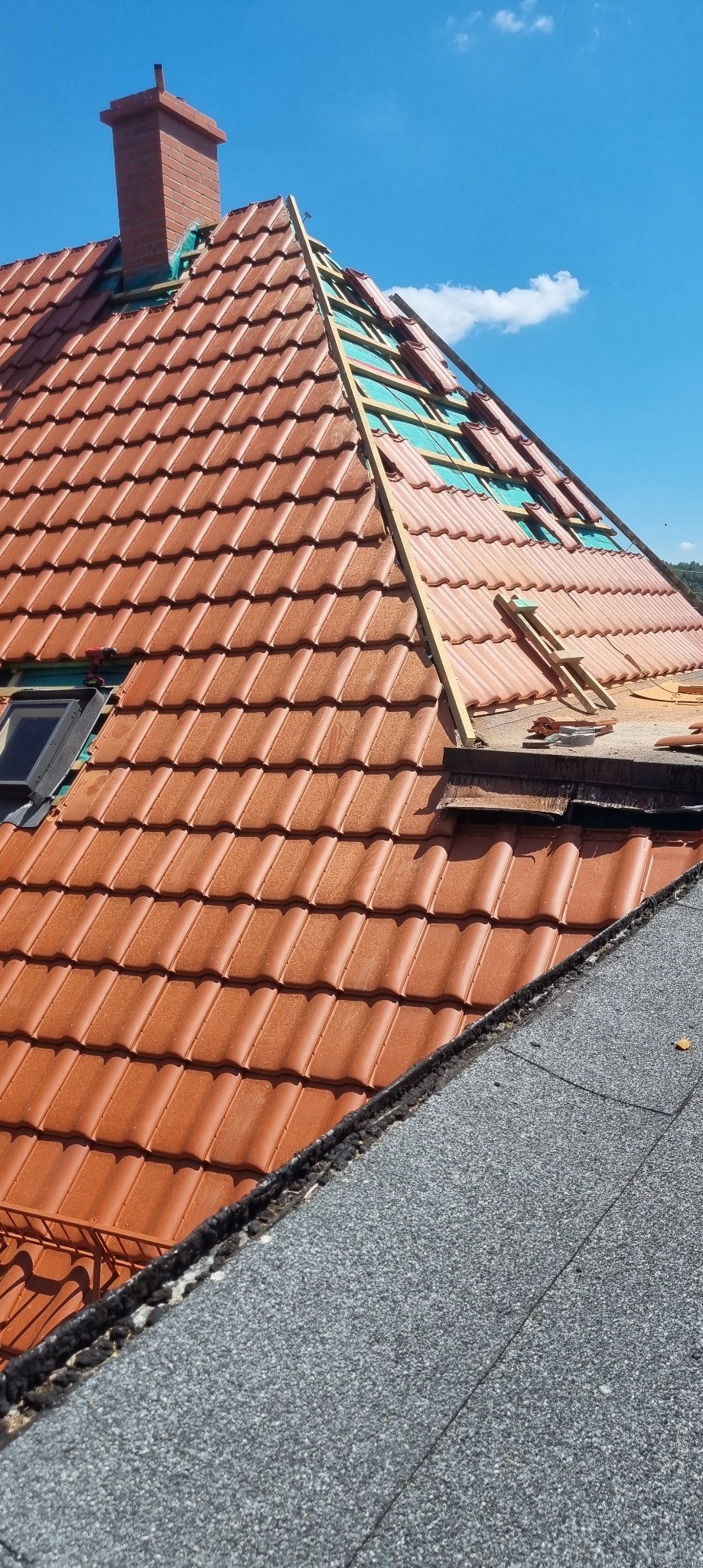 Częściowo zdjęte dachówki z widoczną membraną i drewnianą konstrukcją więźby dachowej oraz komin w tle, fragment dachu pokryty papą.
