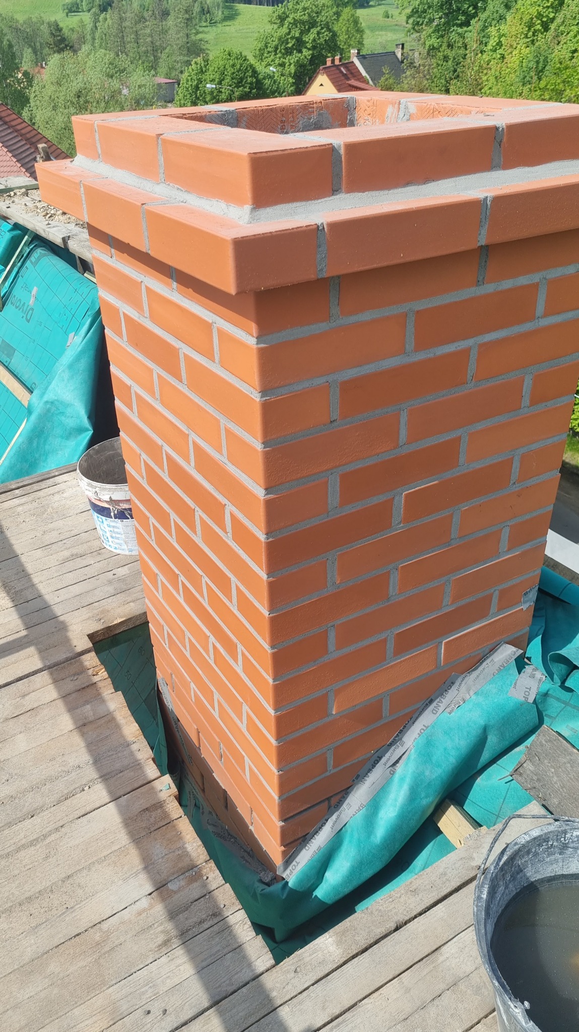 Wzniesiony z cegły komin na dachu, zabezpieczony membraną dachową, widoczne narzędzia i wiadro z zaprawą.