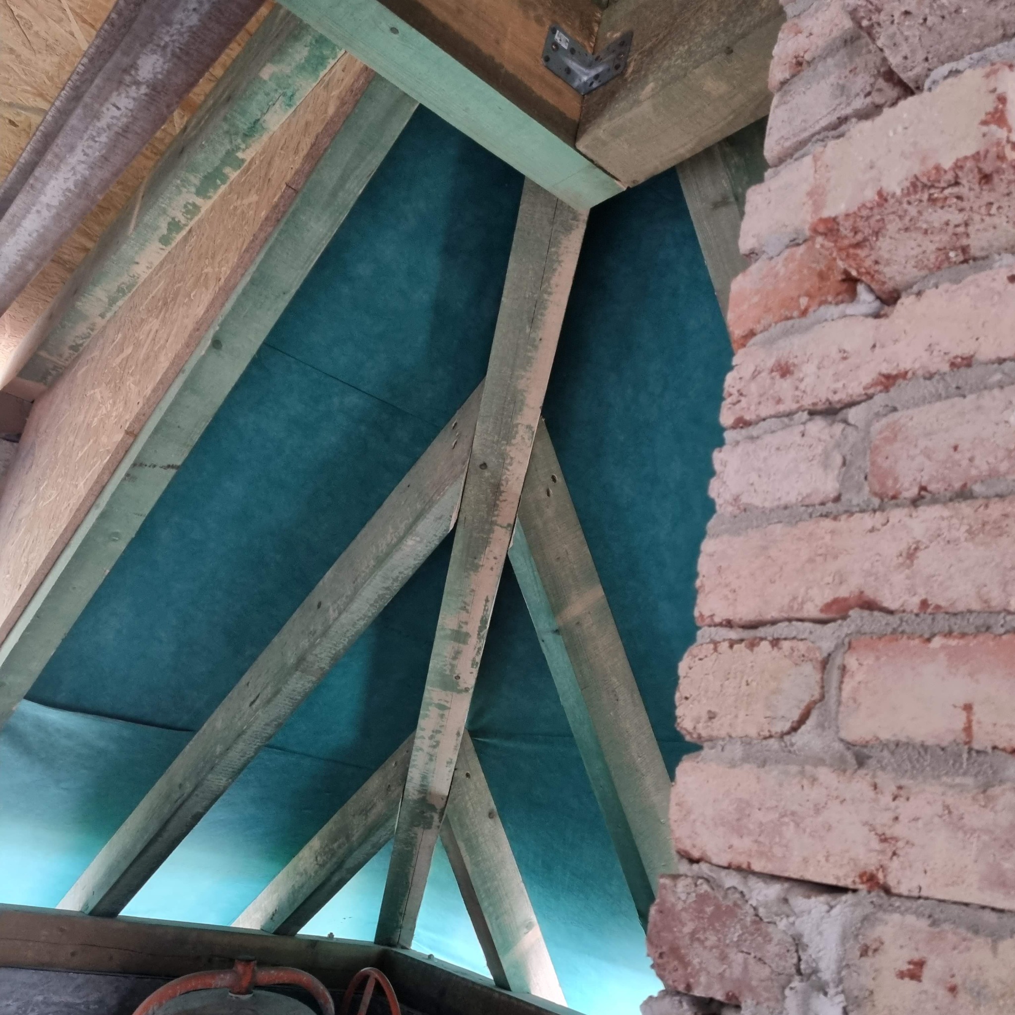 Wnętrze dachu z widoczną konstrukcją drewnianą, membraną dachową i fragmentem murowanej ściany z cegły.