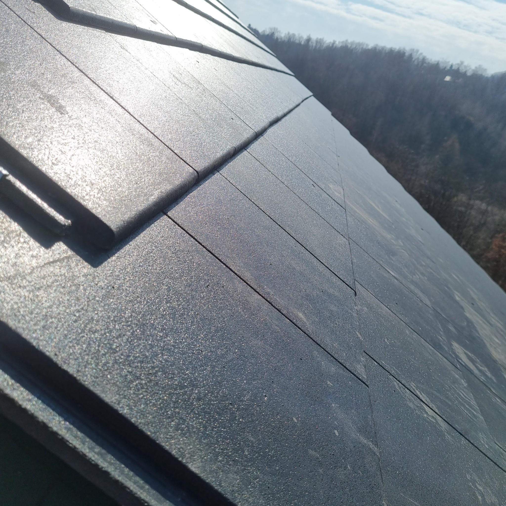 Nowy dach pokryty grafitową dachówką modułową, widok pod kątem w słoneczny dzień, w tle zalesione wzgórza.