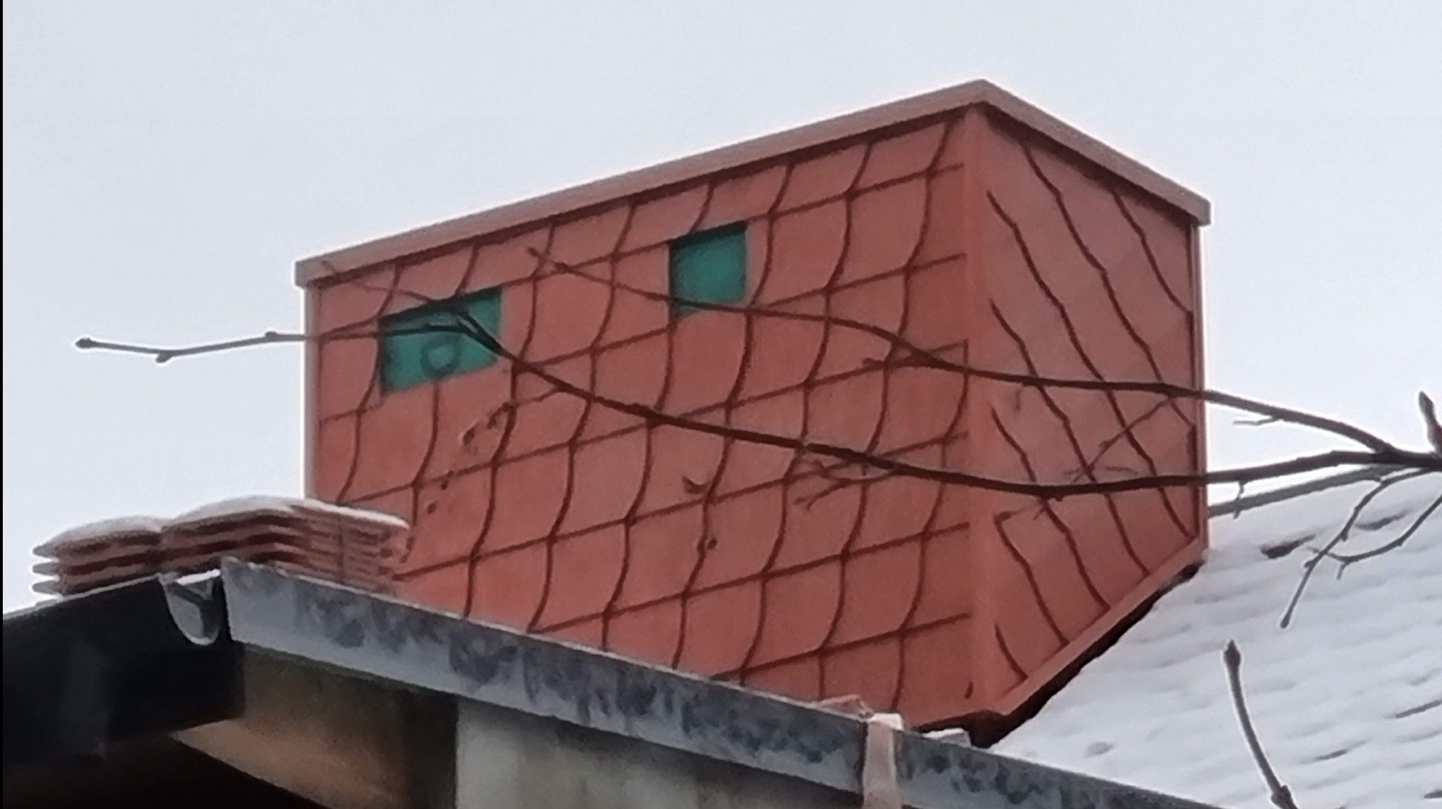 Obróbka komina blachą w kolorze ceglastym, z widocznymi elementami wentylacyjnymi, na tle zimowego krajobrazu.