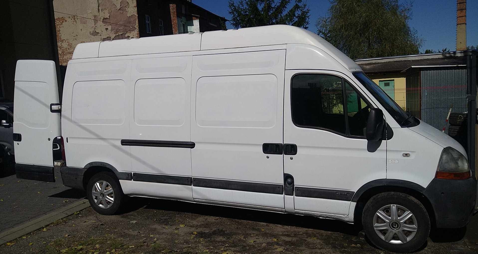 Biały samochód dostawczy z otwartymi tylnymi drzwiami, zaparkowany na zewnątrz budynku, widoczny z boku.