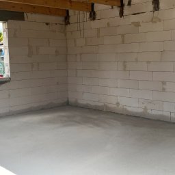 GREKBUD USŁUGI REMONTOWO-BUDOWLANE KACPER DOMŻALSKI - Surowy stan wnętrza domu murowanego z bloczków, widoczne drewniane belki stropowe, w oknie widoczne butelki z napojami.