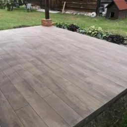 Ułożony taras z płytek imitujących drewno na zewnątrz, z obrzeżem, widoczne rośliny ozdobne, trawnik i elementy architektury ogrodowej w tle.