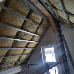 GREKBUD USŁUGI REMONTOWO-BUDOWLANE KACPER DOMŻALSKI - Poddasze w trakcie ocieplania wełną mineralną, widoczne metalowe profile konstrukcyjne i fragment rusztowania z desek, okno w ścianie szczytowej.