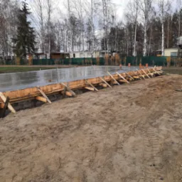 Wylany fundament z drewnianym szalunkiem i pomarańczowymi rurami wentylacyjnymi, widok z poziomu gruntu na tle drzew i zabudowań.