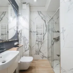 Jasna łazienka z marmurowymi płytkami na ścianach, drewnopodobną podłogą, owalnym lustrem i szklaną kabiną prysznicową.