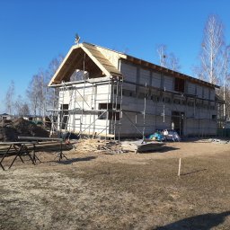 GREKBUD USŁUGI REMONTOWO-BUDOWLANE KACPER DOMŻALSKI - Budowa domu murowanego w trakcie realizacji: ściany z bloczków, drewniana konstrukcja dachu, rusztowania, plac budowy z ziemią i drewnem, błękitne niebo.