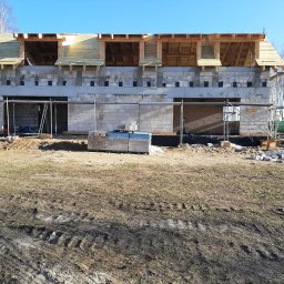 GREKBUD USŁUGI REMONTOWO-BUDOWLANE KACPER DOMŻALSKI - Surowy stan domu z bloczków betonowych z drewnianą konstrukcją dachu na etapie budowy, widoczne rusztowania, palety z materiałami budowlanymi i ślady opon na ziemi