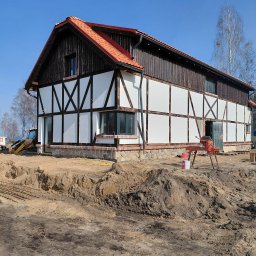 GREKBUD USŁUGI REMONTOWO-BUDOWLANE KACPER DOMŻALSKI - Budowa domu w stylu szachulcowym z czerwoną dachówką. Widoczny wykop pod fundamenty, żółta koparka i narzędzia budowlane. Słoneczny dzień, błękitne niebo.