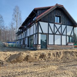 GREKBUD USŁUGI REMONTOWO-BUDOWLANE KACPER DOMŻALSKI - Budowa domu w stylu szachulcowym: widoczne rusztowania, fundamenty z kamienia i zbrojenie w wykopie. Jasne niebo, drzewa w tle. Dom w trakcie budowy.