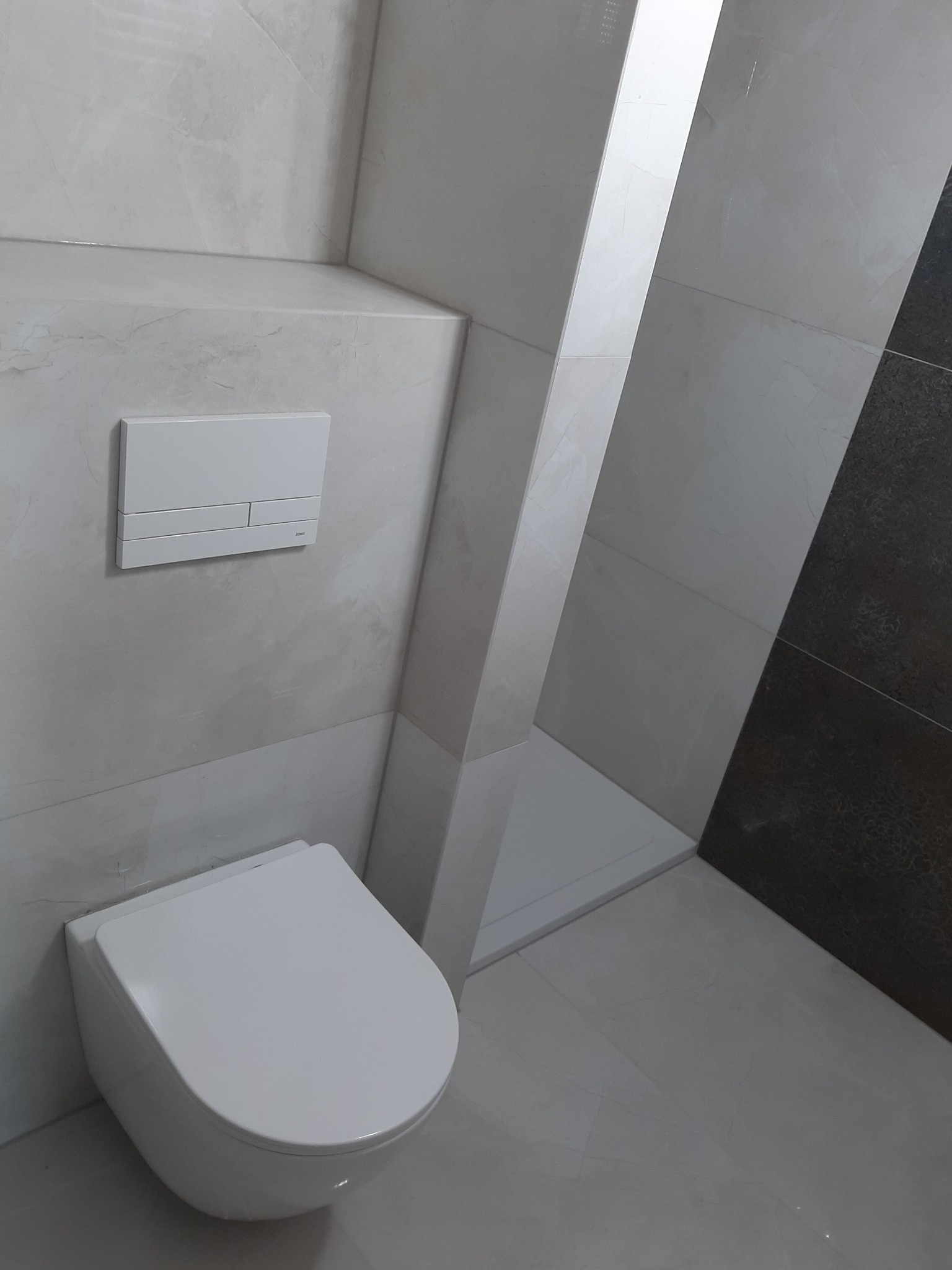 Podwieszana miska WC z przyciskiem spłukującym na marmurowej ścianie, wykończona w jasnych płytkach z ciemnym akcentem.