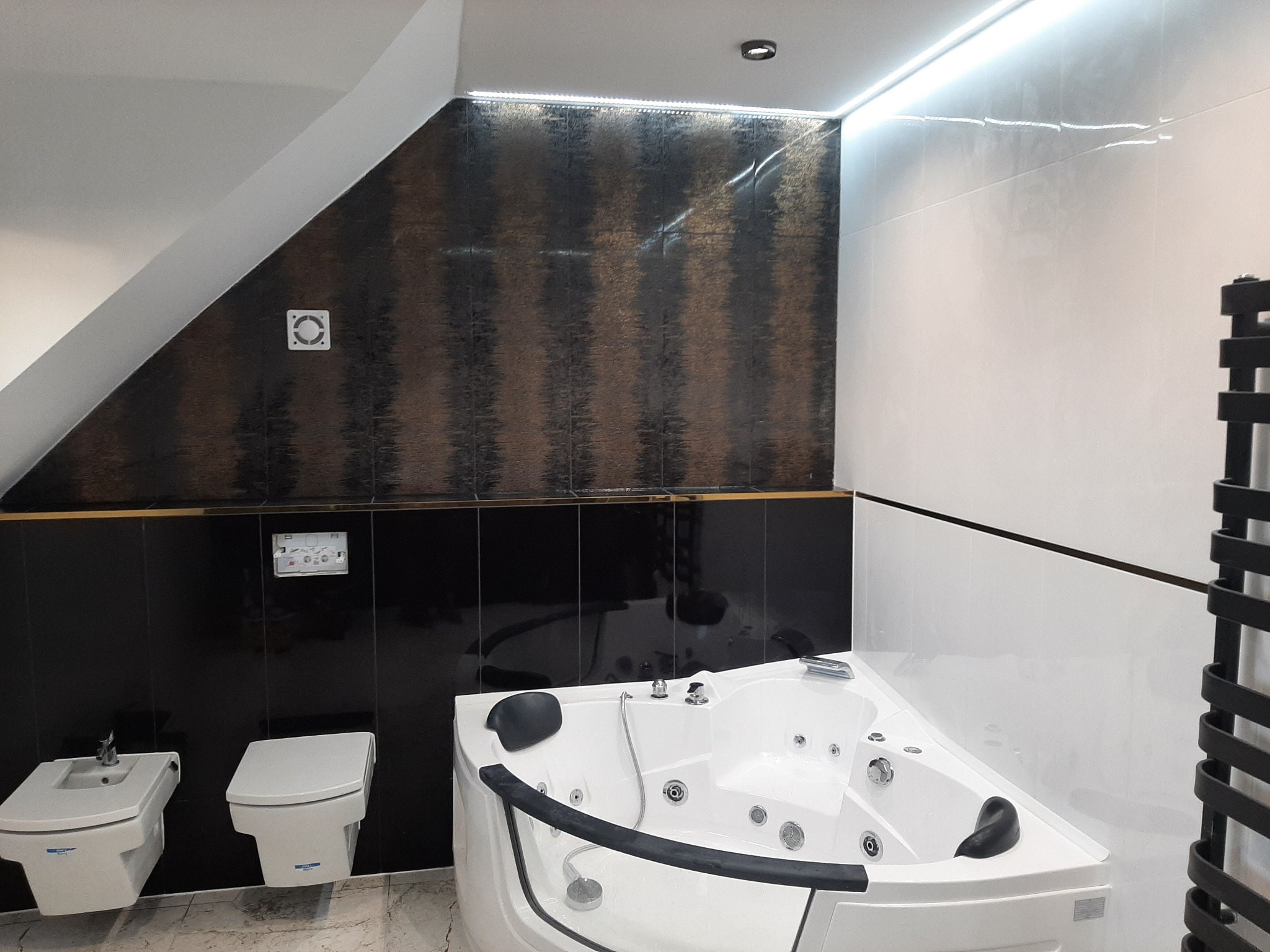 Nowoczesna łazienka na poddaszu z białą wanną z hydromasażem, bidetem i toaletą w zabudowie, wykończona czarno-złotymi i białymi płytkami ceramicznymi oraz oświetleniem LED.