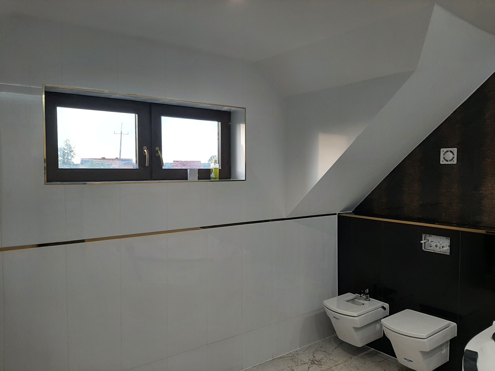 Nowoczesna łazienka na poddaszu z białymi i czarnymi płytkami, armaturą sanitarną w geometrycznym stylu i oknem z widokiem na zewnątrz.