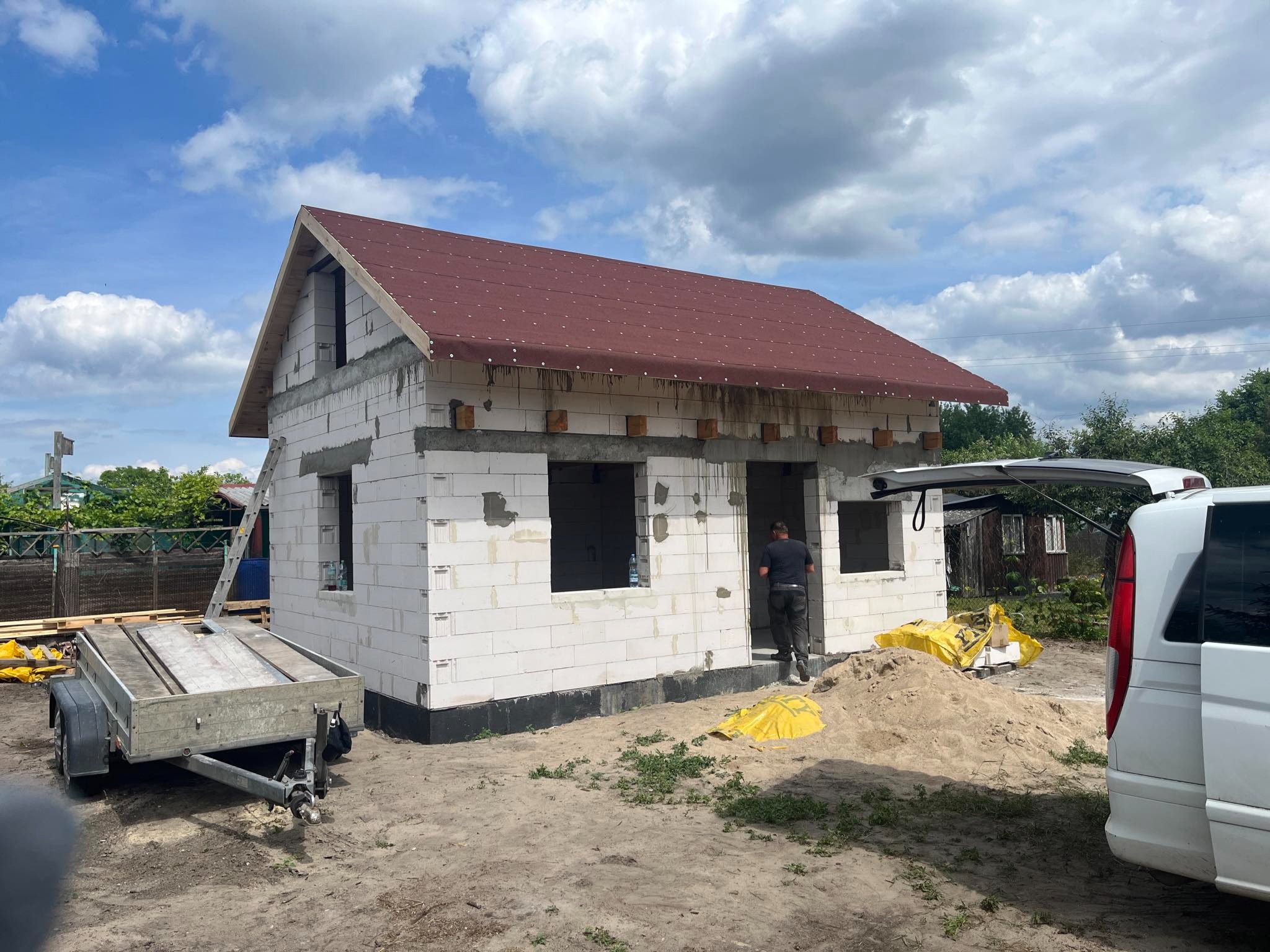 Surowy stan domu z bloczków w Pisz, z prowizorycznym dachem z papy, widoczny przyczepka i częściowo samochód dostawczy na działce.