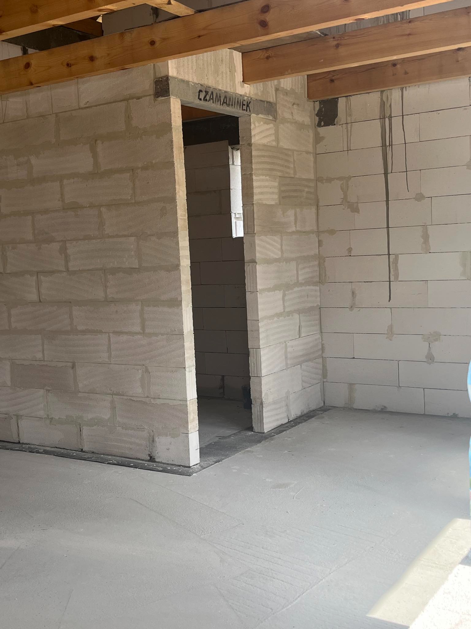 Surowe wnętrze domu w trakcie budowy z widocznymi ścianami z betonu komórkowego, drewnianymi belkami stropowymi i wylewką podłogową, napis 'CZAMANINEK' na nadprożu.