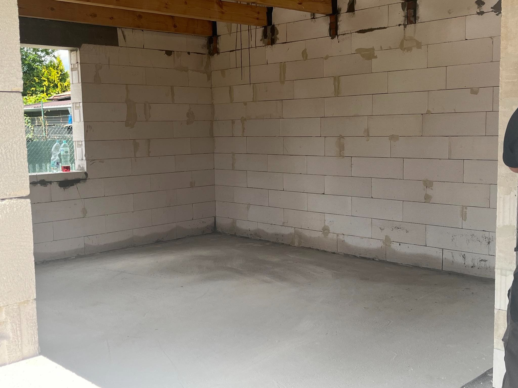 Surowy stan wnętrza domu murowanego z bloczków, widoczne drewniane belki stropowe, w oknie widoczne butelki z napojami.