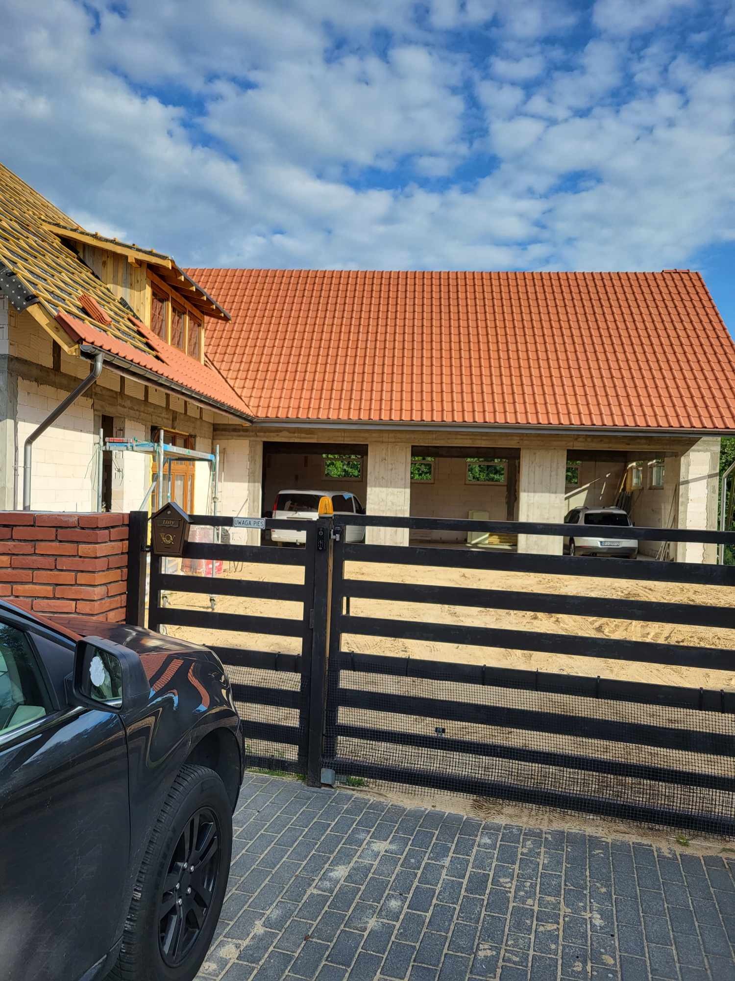 Budynek w trakcie budowy z czerwoną dachówką, widoczny przez czarny płot z furtką, obok zaparkowany samochód. Część budynku w stanie surowym otwartym, z widocznym drewnianym szkieletem dachu. W tle...