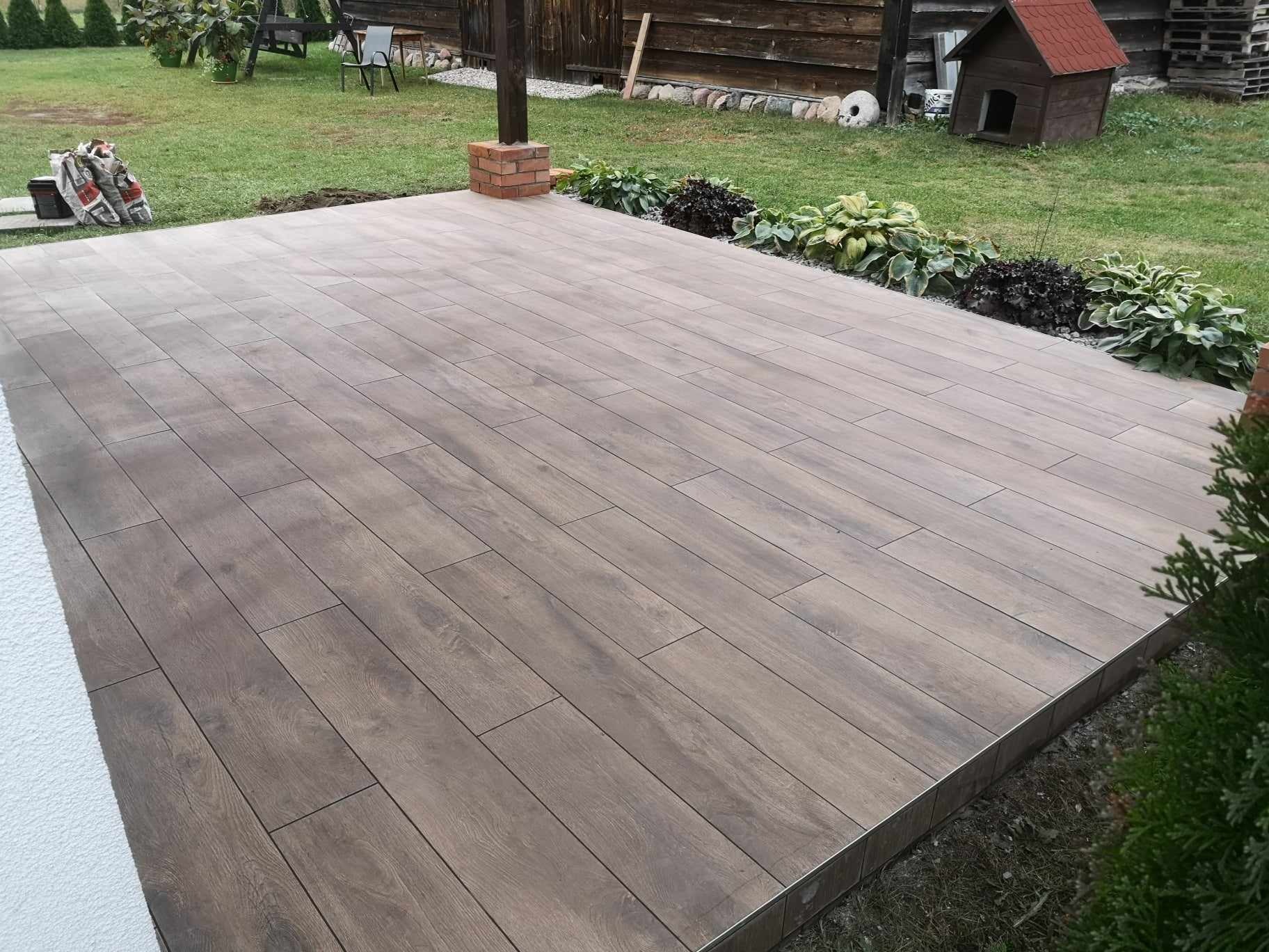 Ułożony taras z płytek imitujących drewno na zewnątrz, z obrzeżem, widoczne rośliny ozdobne, trawnik i elementy architektury ogrodowej w tle.
