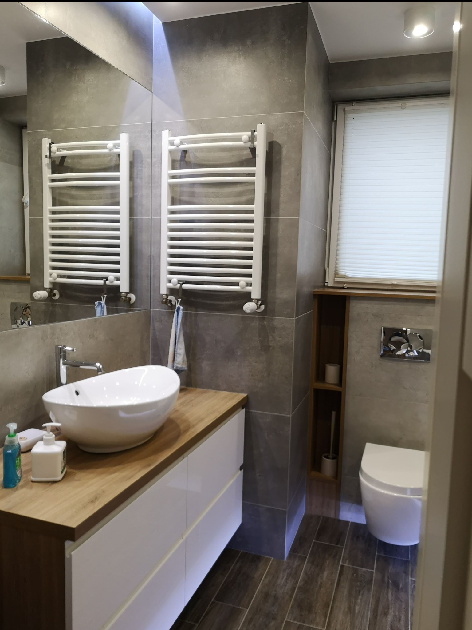 Nowoczesna łazienka z szarymi płytkami, drewnianym blatem pod umywalką nablatową, białymi szafkami i grzejnikiem drabinkowym, widok na toaletę i wnękę z półkami.
