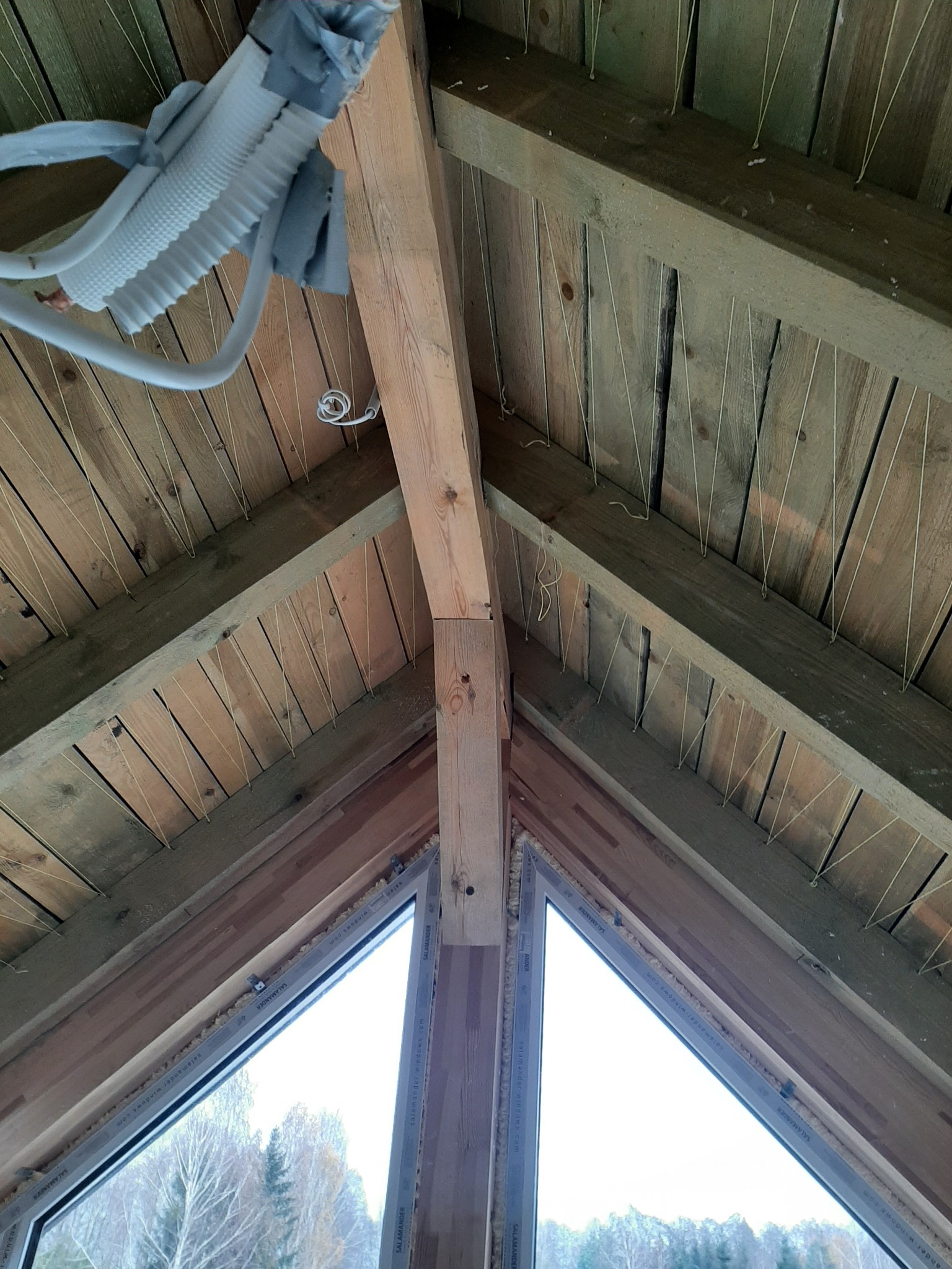 Wnętrze domu w trakcie budowy z widoczną drewnianą konstrukcją dachu, oknami dachowymi i instalacją wentylacyjną, zabezpieczoną folią. Widoczne sznurki przymocowane do drewnianych belek.