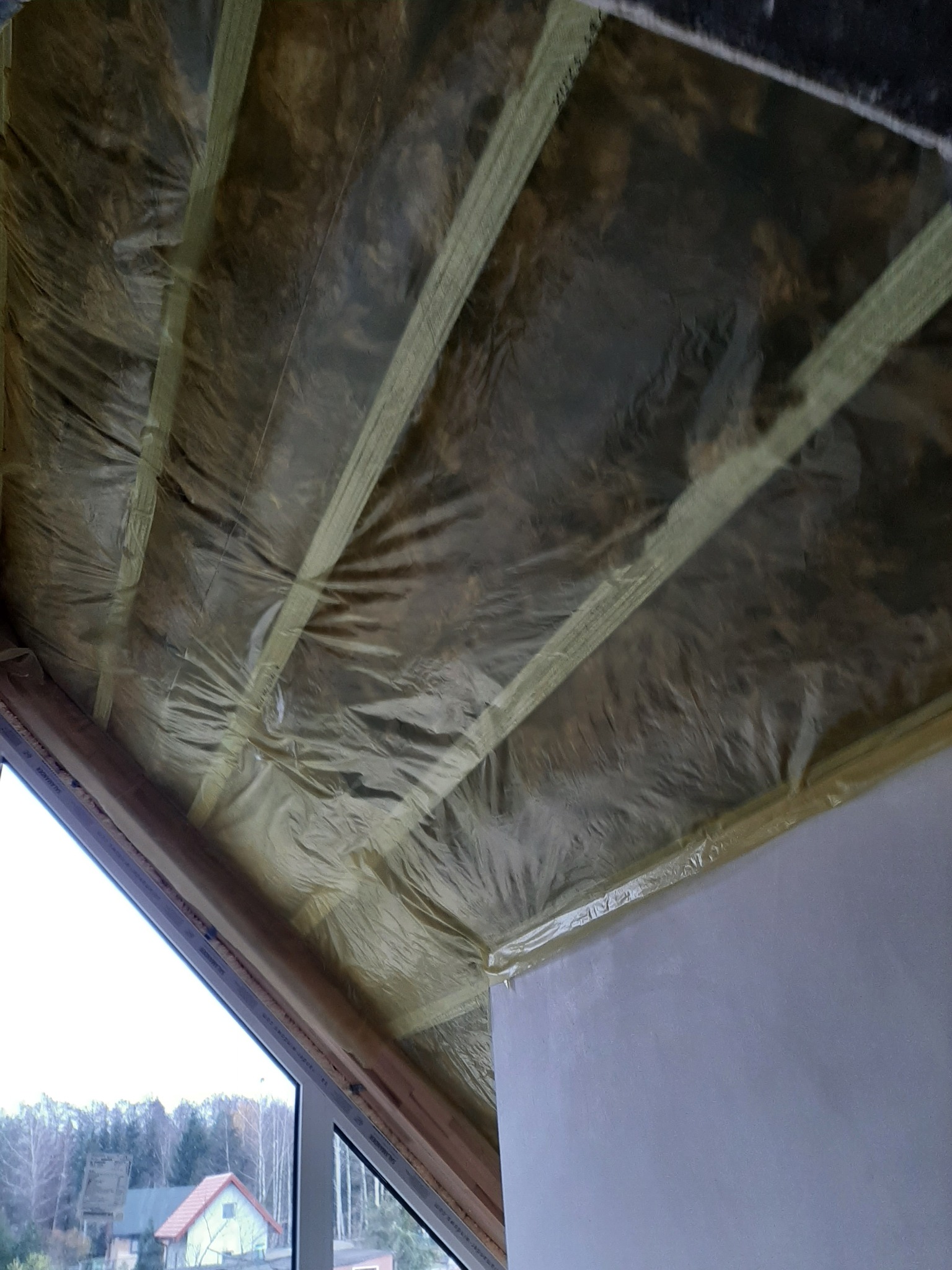 Izolacja poddasza wełną mineralną zabezpieczoną folią paroizolacyjną, widoczne drewniane krokwie i fragment okna z widokiem na krajobraz.