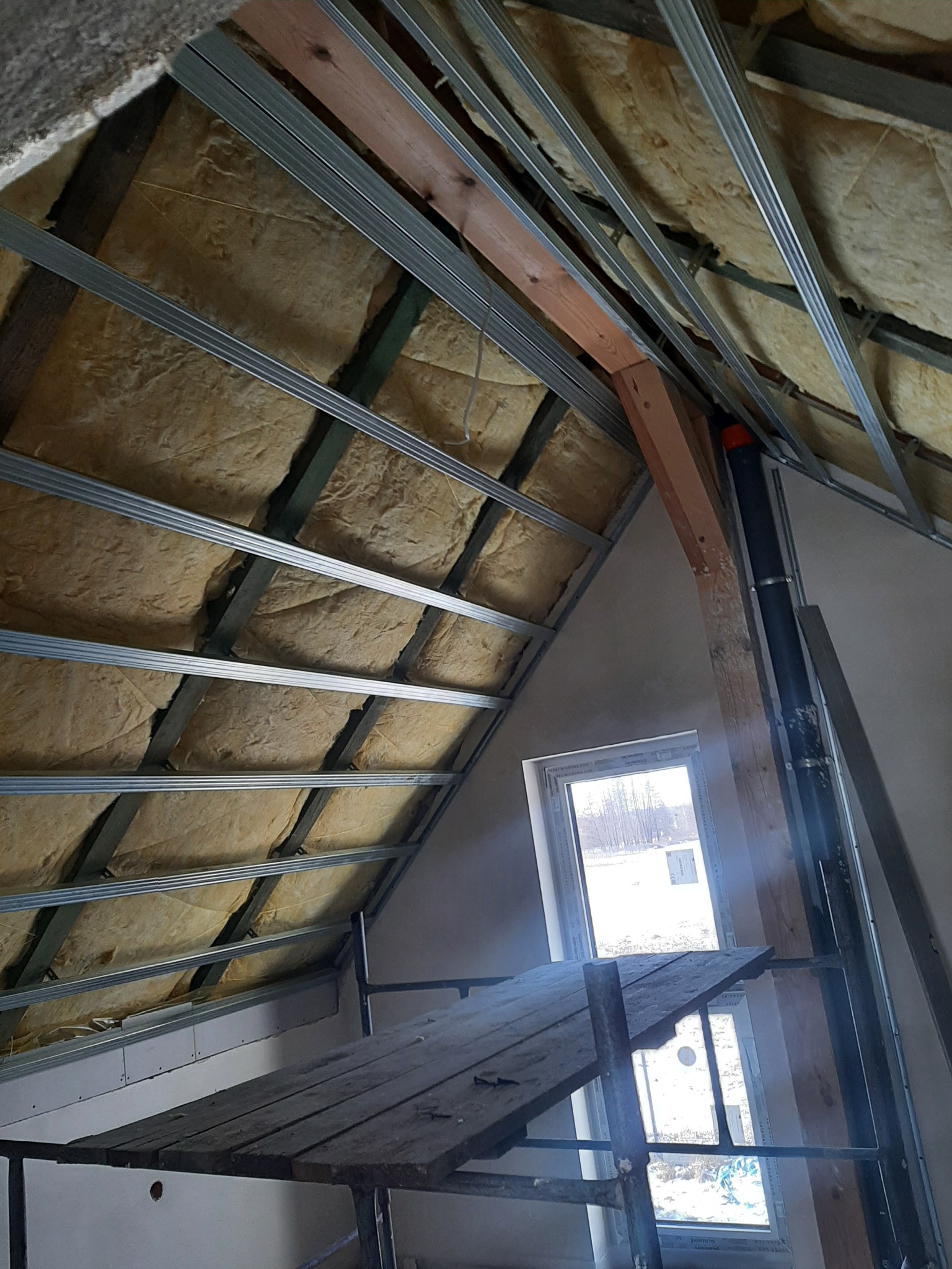 Poddasze w trakcie ocieplania wełną mineralną, widoczne metalowe profile konstrukcyjne i fragment rusztowania z desek, okno w ścianie szczytowej.