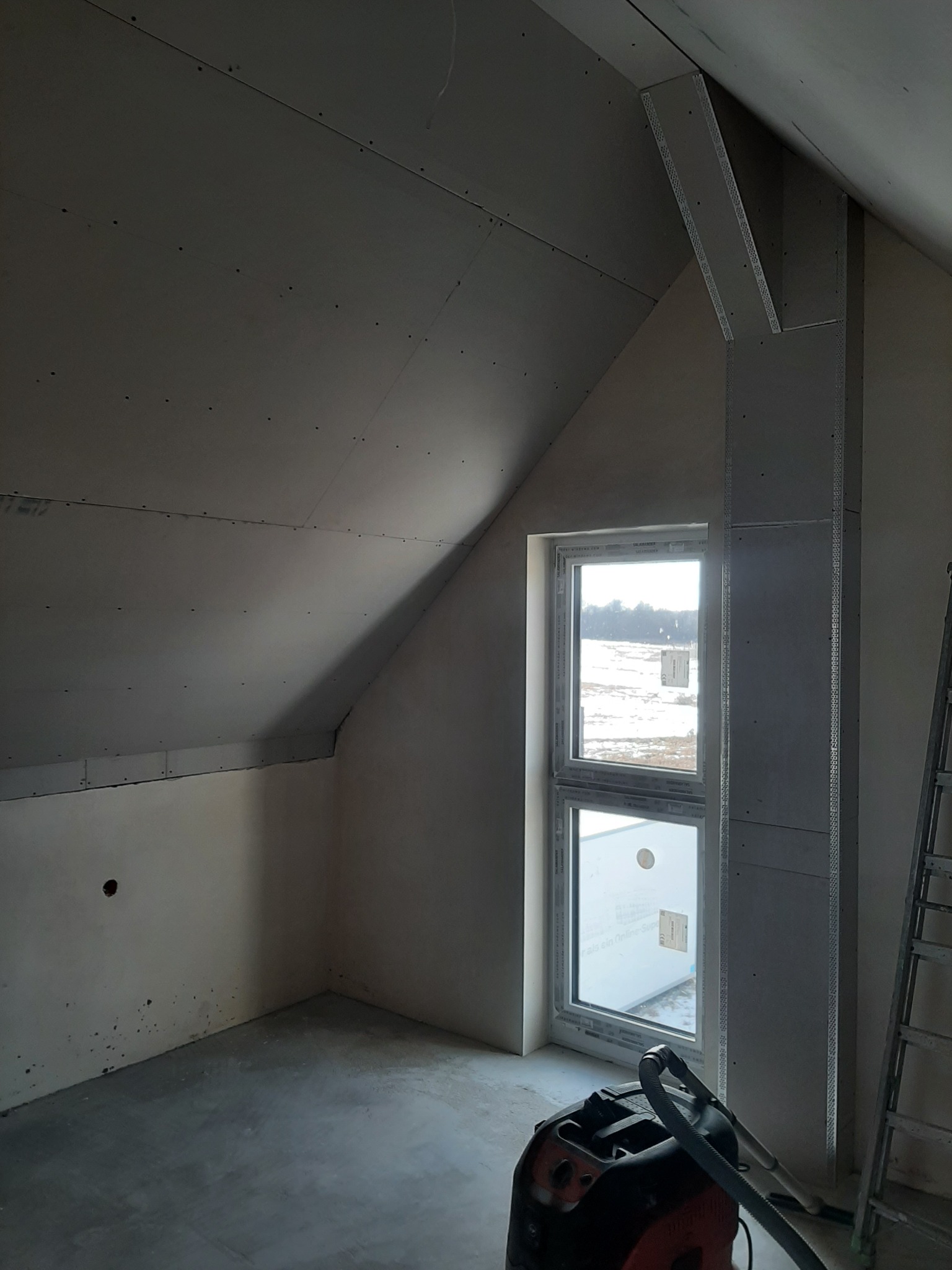 Poddasze w trakcie prac wykończeniowych: zamontowane płyty gipsowo-kartonowe na skosach i ścianach, widoczne profile mocujące, okno z widokiem na zaśnieżone pole, odkurzacz przemysłowy na podłodze,...