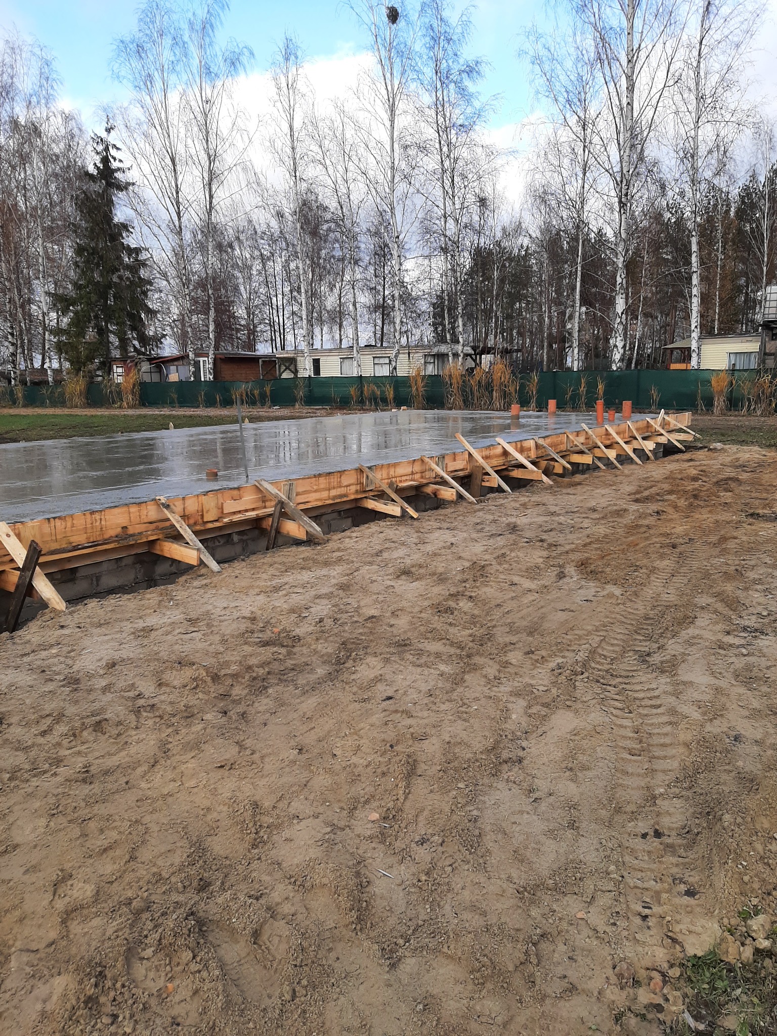 Wylany fundament z drewnianym szalunkiem i pomarańczowymi rurami wentylacyjnymi, widok z poziomu gruntu na tle drzew i zabudowań.