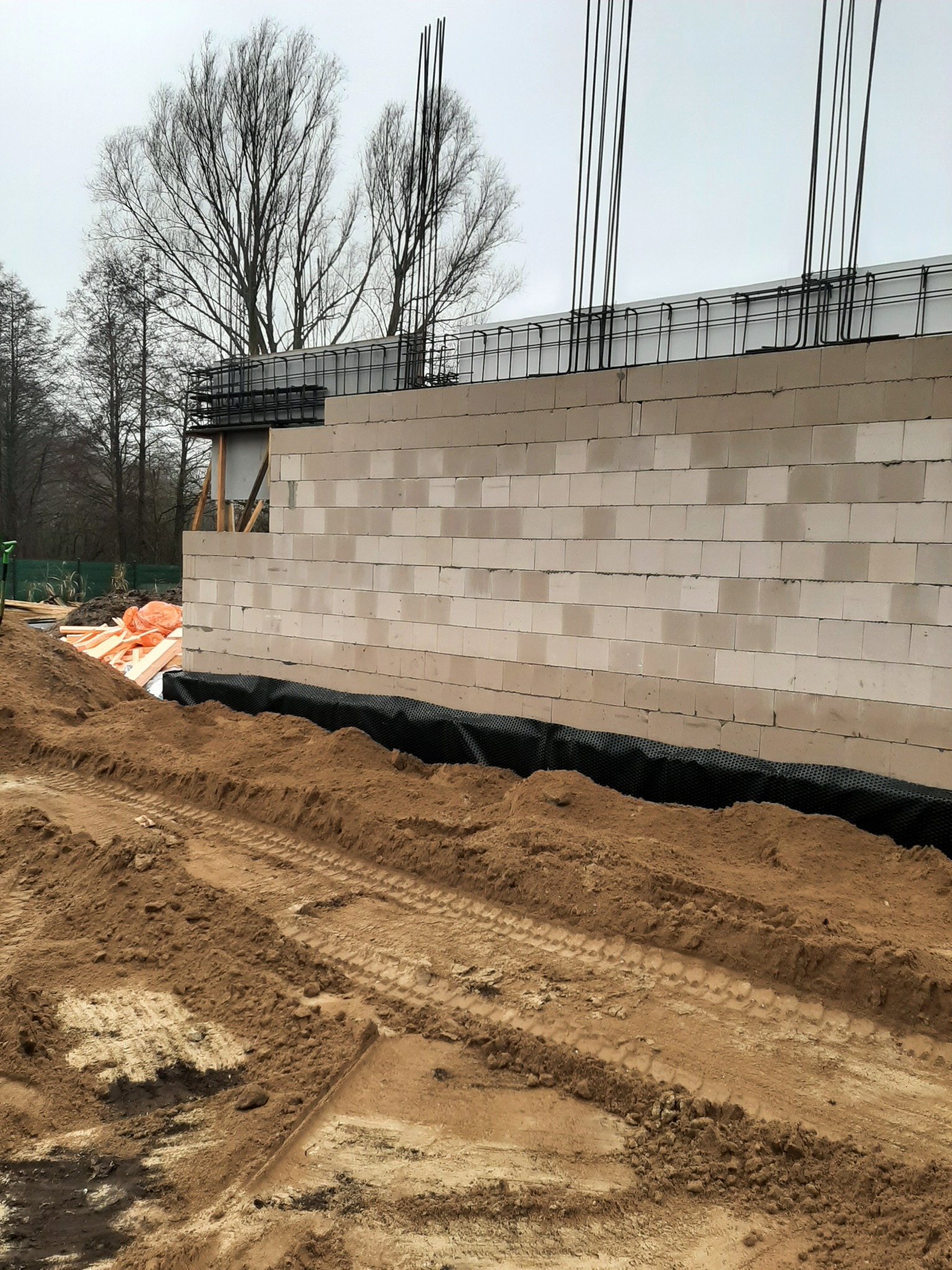 Wznoszone ściany domu z betonu komórkowego z widocznym zbrojeniem i izolacją fundamentów, teren budowy z ziemią i śladami opon.