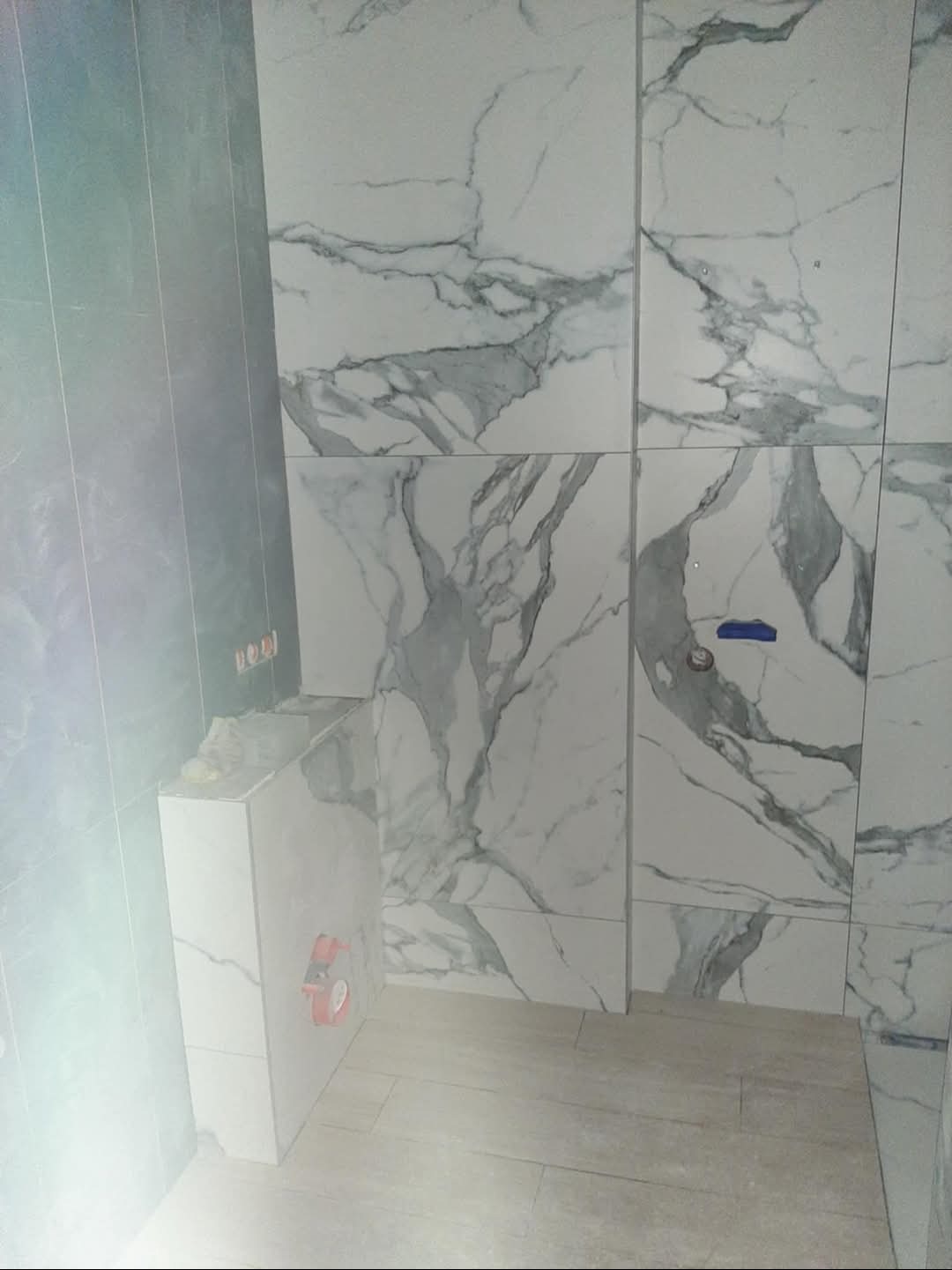 Wnętrze łazienki w trakcie remontu, widoczne płytki imitujące marmur na ścianach i obudowie stelaża podtynkowego, częściowo ułożone panele podłogowe, oraz wystające przewody elektryczne.