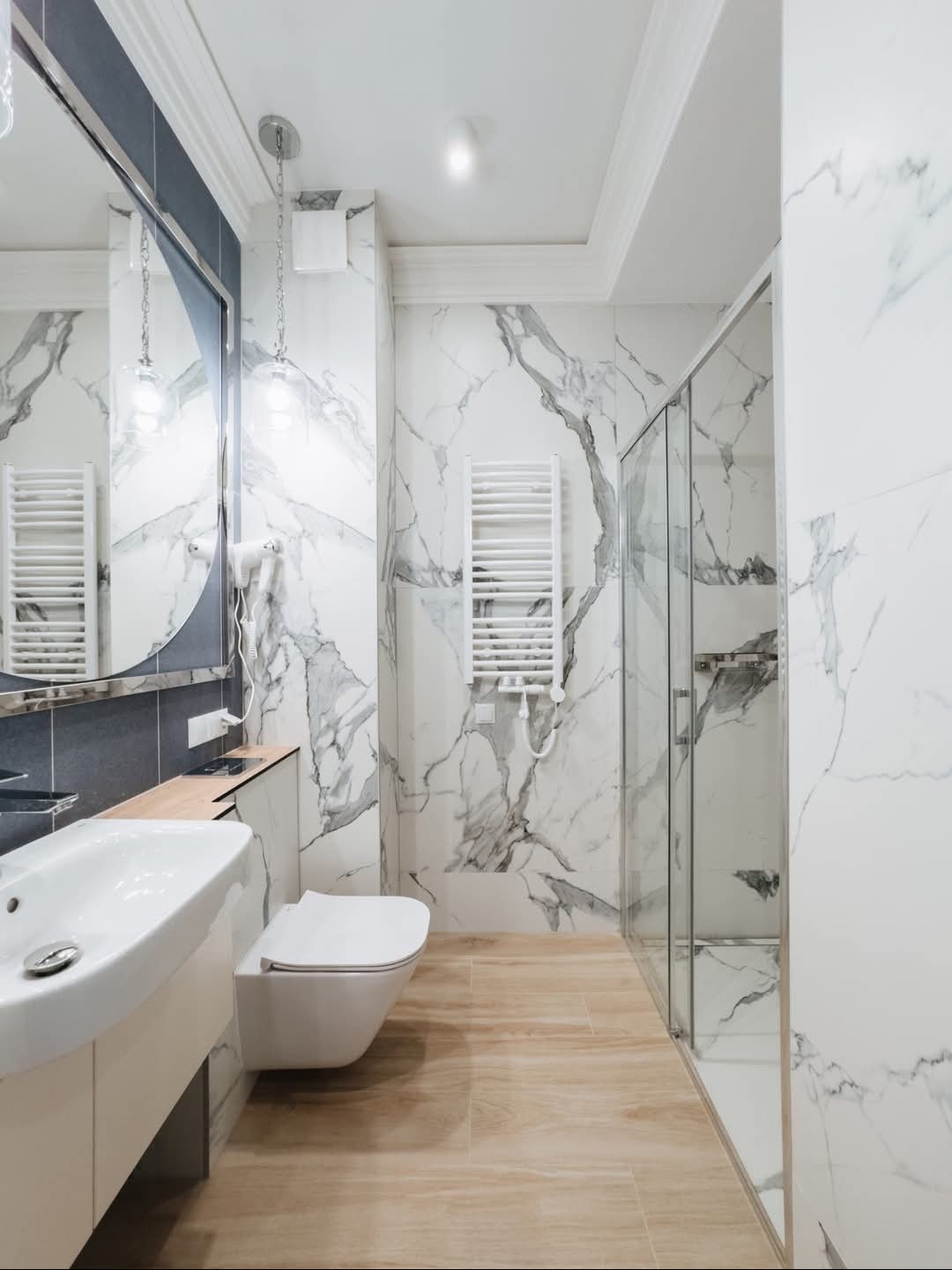 Jasna łazienka z marmurowymi płytkami na ścianach, drewnopodobną podłogą, owalnym lustrem i szklaną kabiną prysznicową.