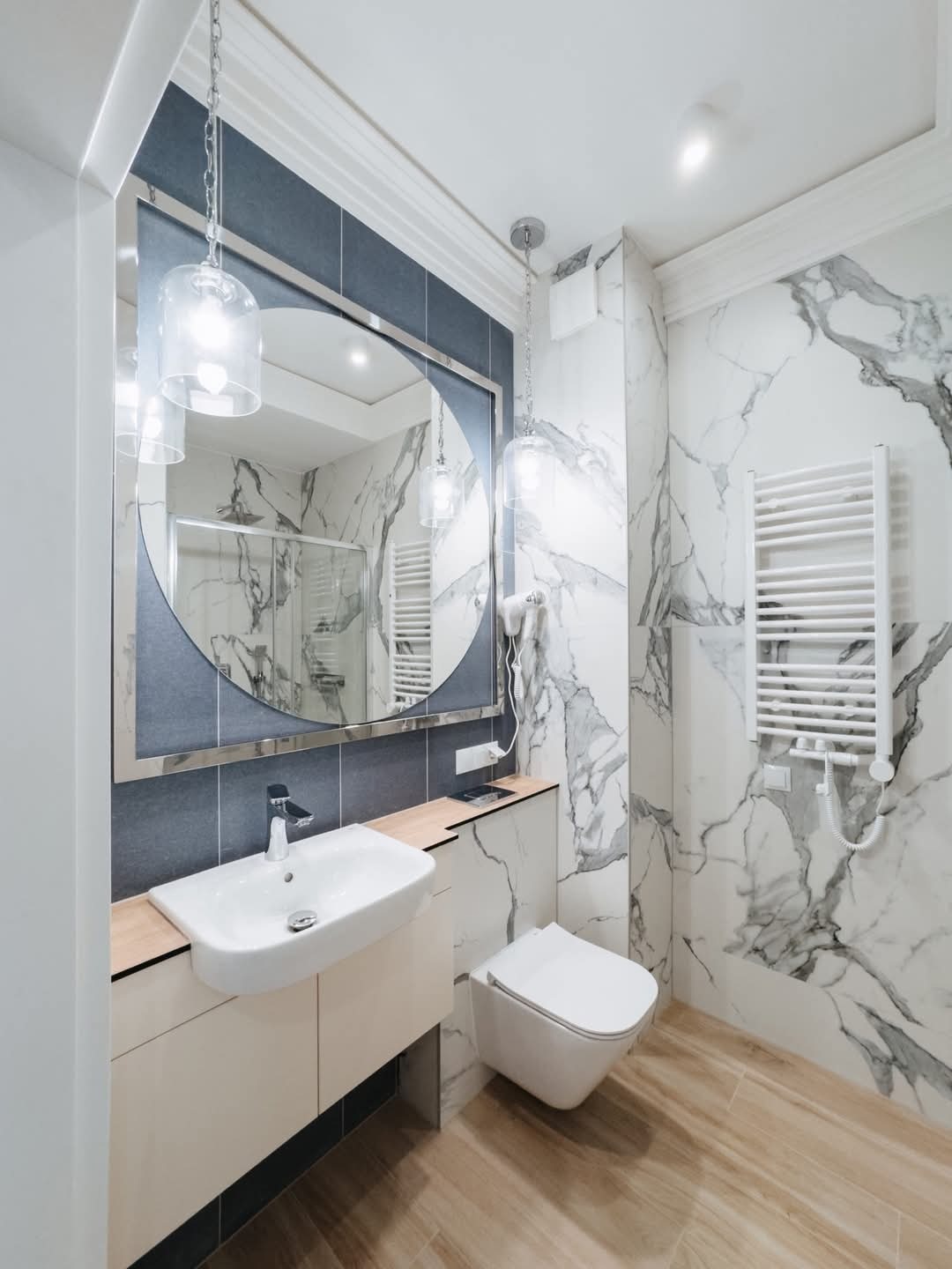 Nowoczesna łazienka z marmurowymi płytkami na ścianach, okrągłym lustrem z podświetleniem, wiszącą umywalką z drewnianym blatem i grzejnikiem drabinkowym.