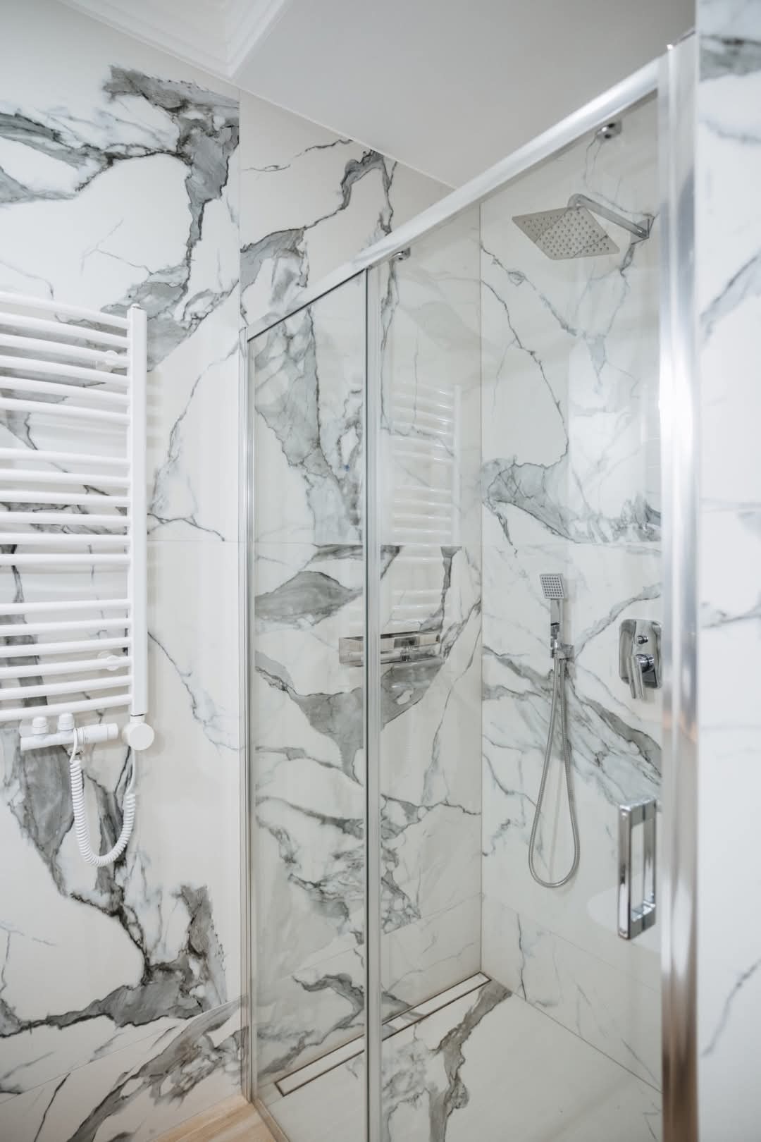 Nowoczesna kabina prysznicowa z przesuwnymi drzwiami, wykończona białymi płytkami imitującymi marmur z szarymi żyłami, widoczny grzejnik drabinkowy i bateria prysznicowa.
