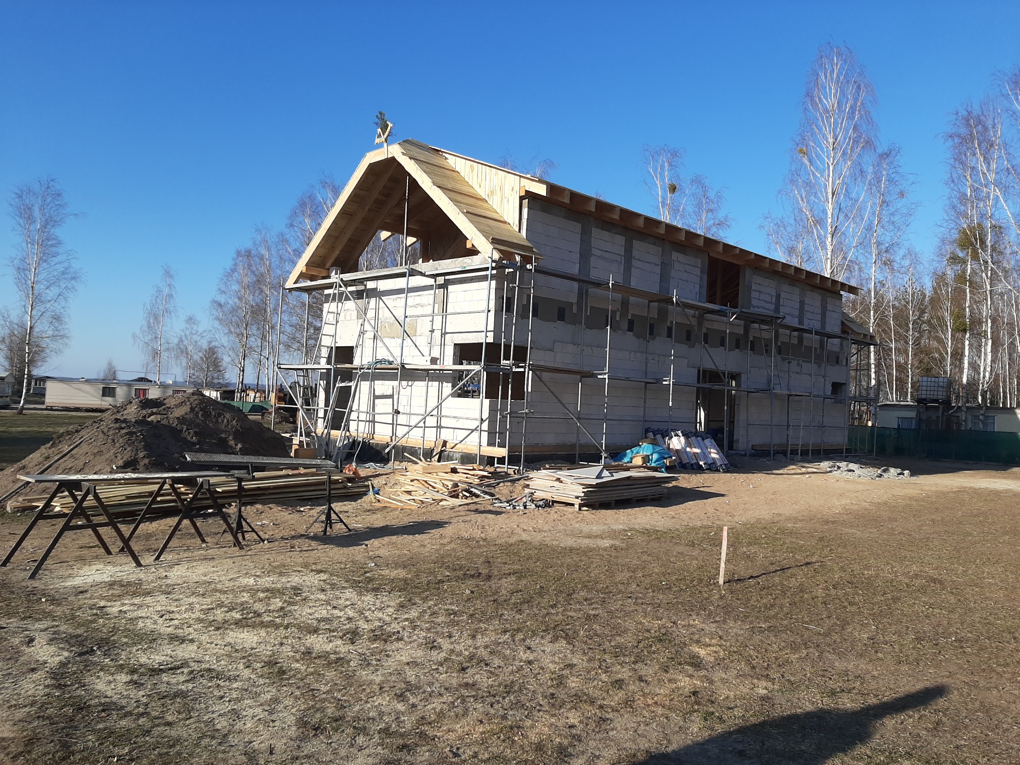 Budowa domu murowanego w trakcie realizacji: ściany z bloczków, drewniana konstrukcja dachu, rusztowania, plac budowy z ziemią i drewnem, błękitne niebo.