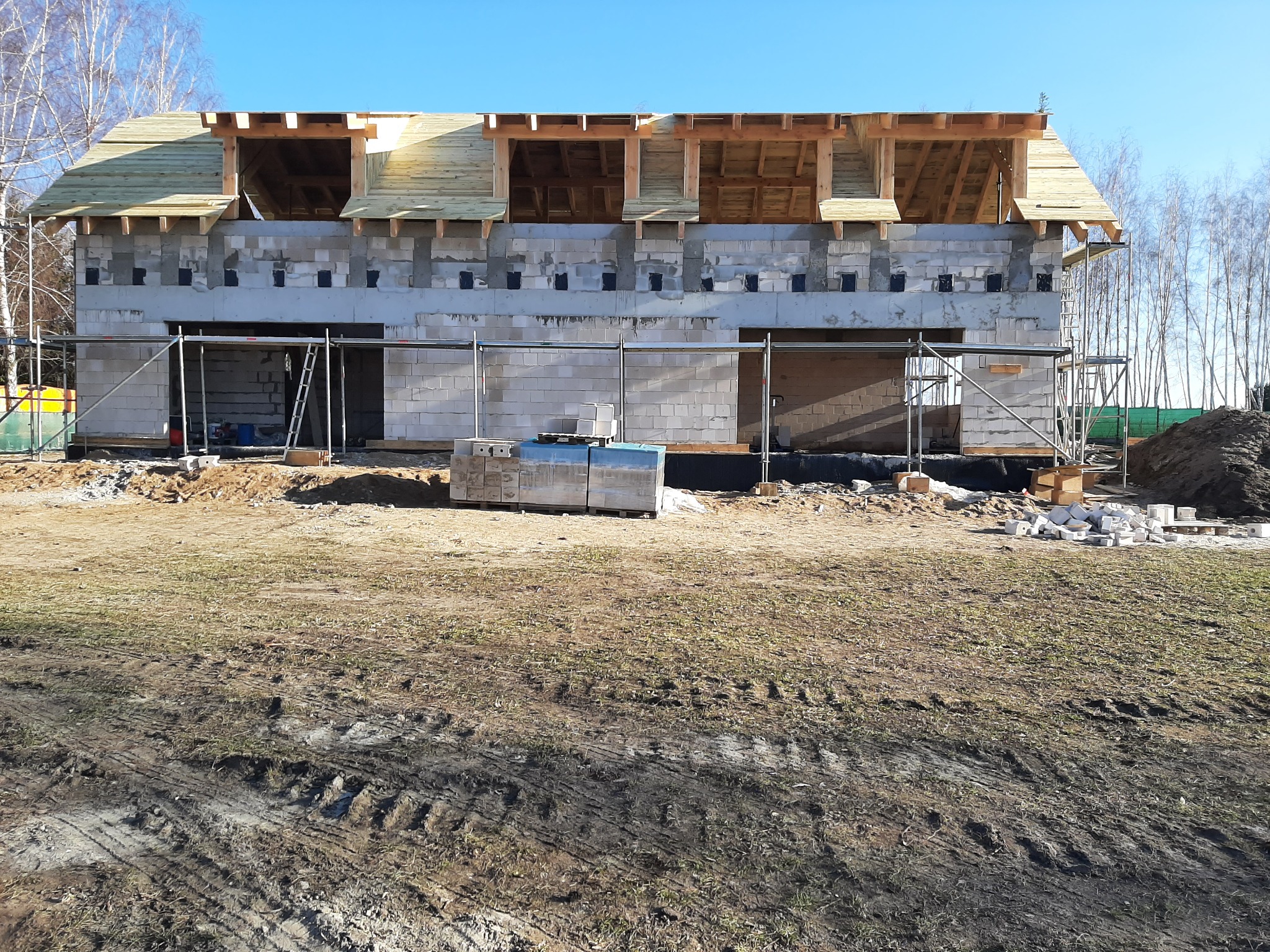 Surowy stan domu z bloczków betonowych z drewnianą konstrukcją dachu na etapie budowy, widoczne rusztowania, palety z materiałami budowlanymi i ślady opon na ziemi