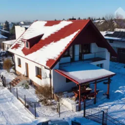 Zimowy widok z góry na dom jednorodzinny z czerwoną dachówką, częściowo pokrytą śniegiem, oraz panele słoneczne na budynku gospodarczym w tle.