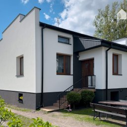 Damian Wesołowski - Odnowiony dom jednorodzinny z białą elewacją i ciemnoszarą podmurówką, brązowe okna, logo firmy w prawym górnym rogu, stół piknikowy z kwiatami na pierwszym planie.