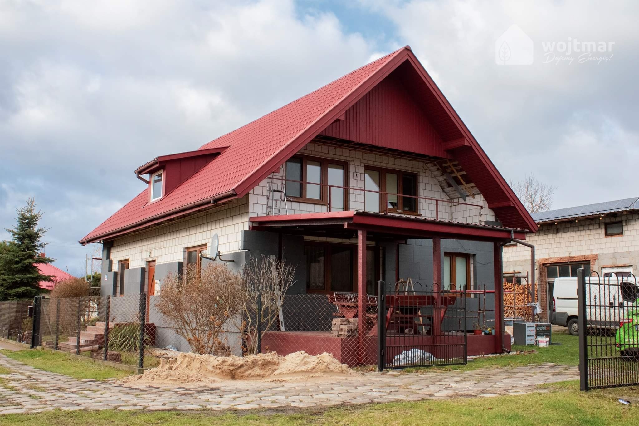Dom jednorodzinny z czerwonym dachem, częściowo ocieplony styropianem, z widocznym fragmentem niezabezpieczonej elewacji i stertą piasku przed ogrodzeniem.